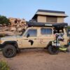 A tan truck with a tent on top parked in the dirt.