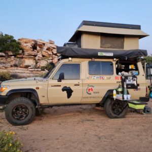 A tan truck with a tent on top parked in the dirt.