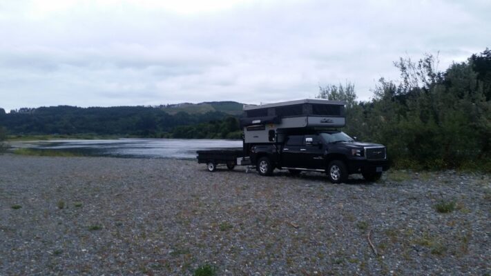 A truck with a camper on the side of a river.